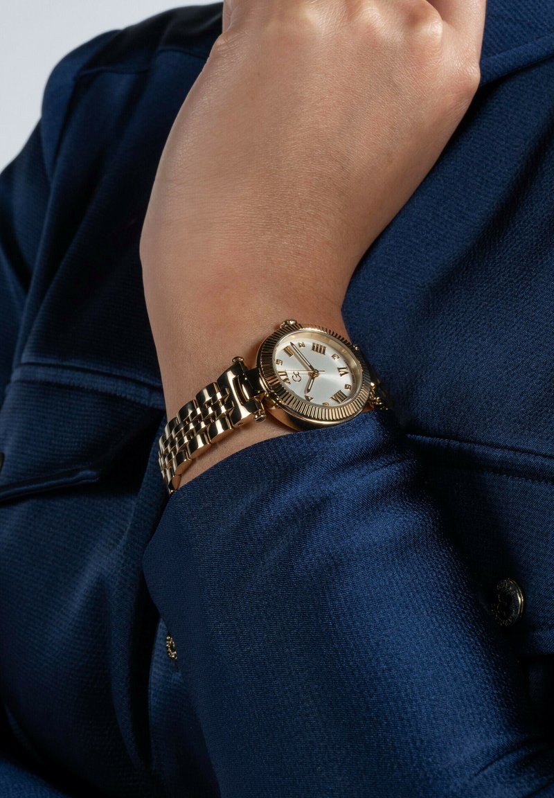 Orologio da polso color oro con quadrante rotondo, dial bianco, numeri romani e bracciale a maglie metalliche, indossato su un polso contro uno sfondo di tessuto blu navy.