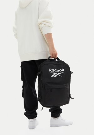 Person hält einen schwarzen Reebok-Rucksack am oberen Griff, trägt einen weißen Hoodie, schwarze Hose und weiße Reebok-Sneaker.