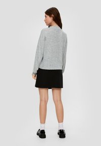 Młoda kobieta stoi, ubrana w szary sweter, czarną spódnicę, białe skarpetki i czarne buty, zwrócona plecami w kierunku jednolitego tła.