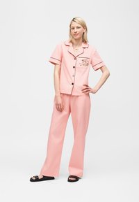 Junge Frau steht mit einer Hand in der Hüfte, trägt einen rosa Pyjama mit Drachenstickerei auf der Tasche und schwarze Pantoletten.