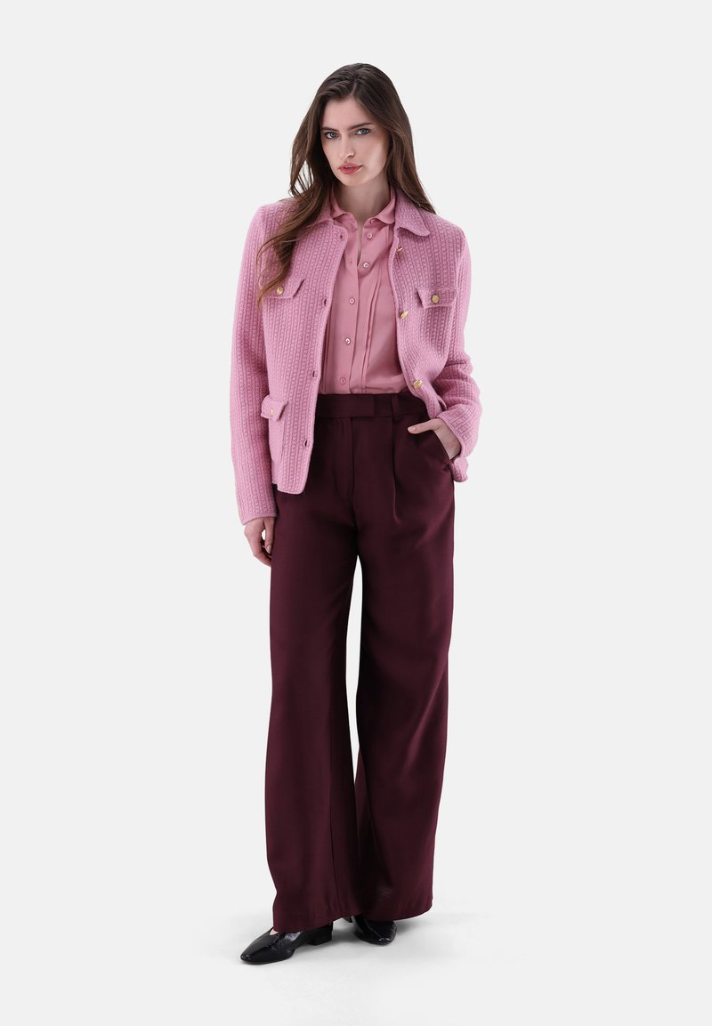 Eine Frau steht da und trägt ein rosa geknöpftes Hemd, eine strukturierte rosa Jacke, weite bordeauxfarbene Hosen und schwarze Schuhe, mit einer Hand in der Tasche.