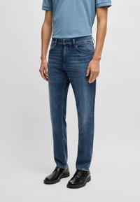 Blue denim jeans with a straight cut, slight fading, five-pocket design, and a zip fly. Paired with black lace-up shoes and a light blue shirt.