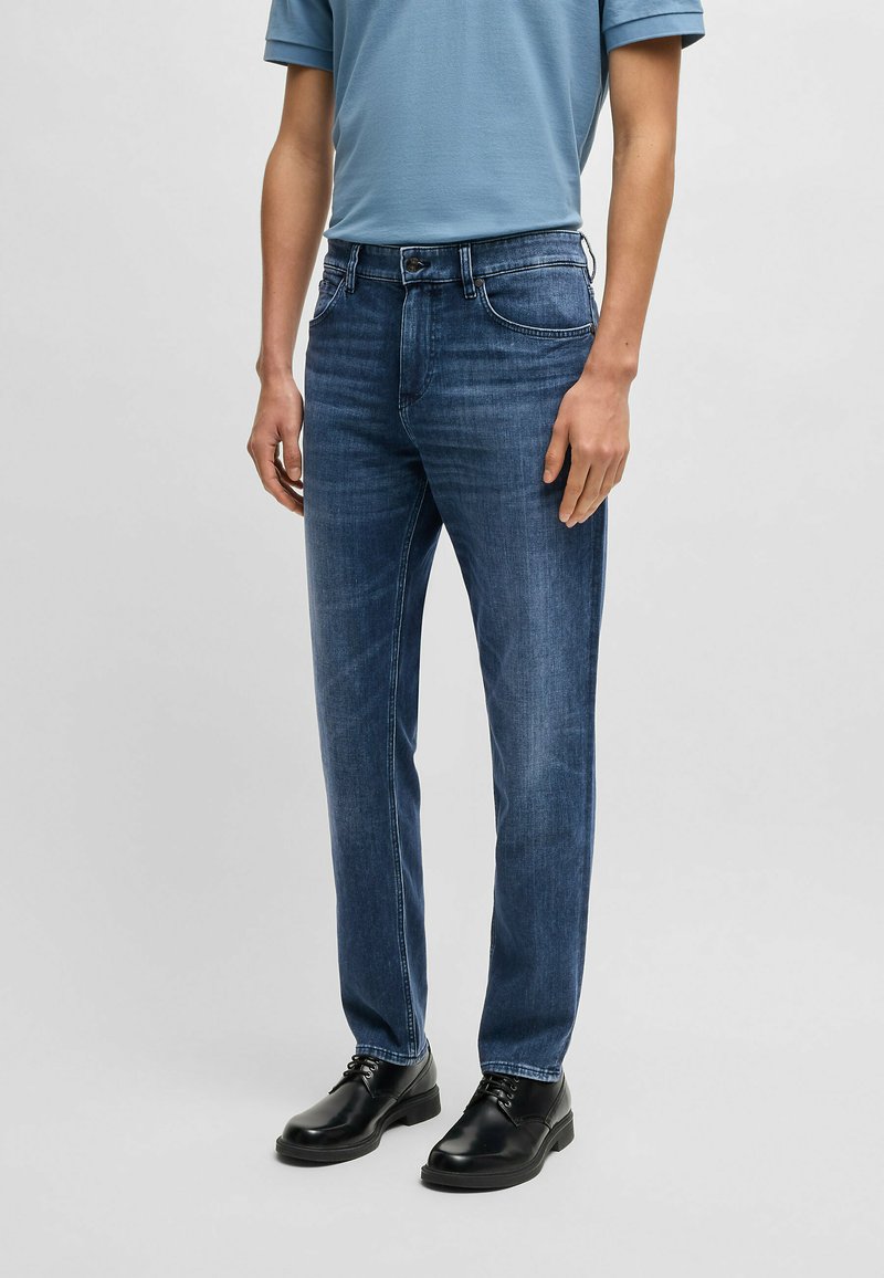 Blue denim jeans with a straight cut, slight fading, five-pocket design, and a zip fly. Paired with black lace-up shoes and a light blue shirt.