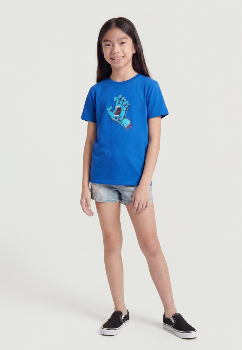 Junges Mädchen lächelt, trägt ein blaues T-Shirt mit einer Grafik einer schreienden blauen Hand, hellblaue Jeansshorts und schwarze Slip-On-Schuhe.