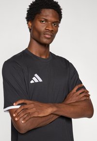 Μαύρο αθλητικό μπλουζάκι με κοντά μανίκια, με λευκό λογότυπο με τρεις ρίγες στο αριστερό στήθος. Λείο υλικό με αντίθετη λευκή επένδυση.