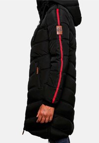 Veste longue noire matelassée avec des bandes rouges sur les manches, une poche zippée à l'avant et un écusson logo sur le haut du bras, portée par une personne debout.