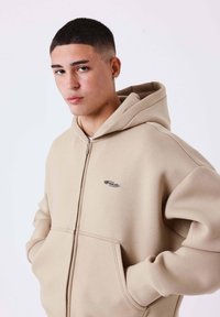 Giovane con capelli corti che indossa una felpa beige con cerniera e le mani nelle tasche, rivolto verso la fotocamera con espressione neutra su sfondo bianco.