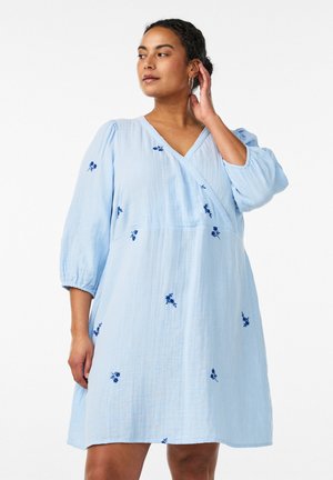 Kvinde i en lyseblå, knælængde kjole med trekvartærmel og små mørkeblå blomsterbroderier, der rører ved sit hår.