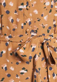 Tissu beige avec imprimé floral en blanc, vert, marine et rose, doté de boutons recouverts de tissu et d'un nœud de ceinture noué.