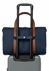 Sac de voyage bleu marine avec des accents en cuir marron, attaché à une valise rigide grise, dotée d'une poignée télescopique et d'une texture lisse.