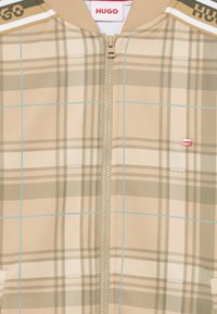 Giacca beige a quadri con zip e dettagli azzurri, presenta un corpo texturizzato, colletto a coste e un piccolo logo rosso sul petto.