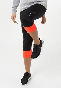Leggings atléticos negros con acentos naranjas en las rodillas, que presentan detalles reflectantes. Combinados con zapatillas de correr negras.
