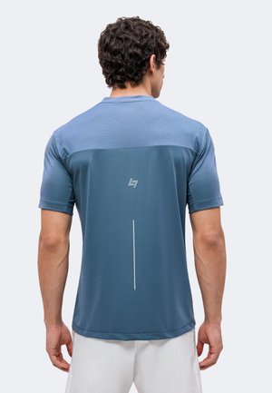 Homme debout de dos, portant un t-shirt de sport bleu à manches courtes et un short blanc sur un fond clair uni.