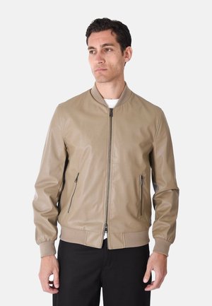 Uomo che indossa una giacca bomber beige con zip, polsini e orlo a coste, pantaloni neri, in piedi su sfondo bianco.