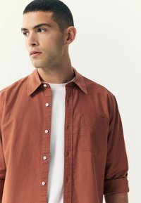 Chemise brune à manches longues avec un col et une poche poitrine, portée sur un T-shirt blanc. Le tissu semble doux avec un design décontracté.