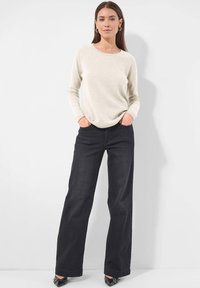 Frau in beige Langarmpullover und hochtaillierten, weit geschnittenen schwarzen Hosen, die mit den Händen in den Taschen steht und schwarze spitze Schuhe trägt.
