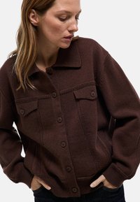 Pull en tricot marron à boutonnière avec un col, doté de deux poches poitrine, de poignets côtelés et d'un motif texturé sur l'ensemble.