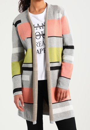 Kvinde iført en lang cardigan med åbent frontstykke med grå, pink, grønne og sorte geometriske striber over en hvid grafisk t-shirt.
