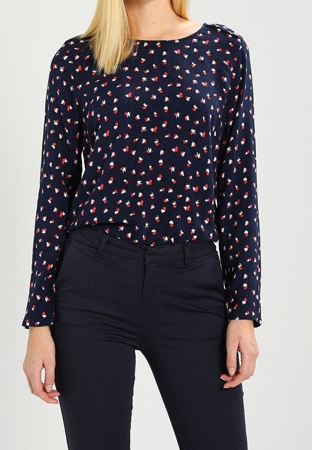Femme portant une blouse bleu marine à manches longues à motifs floraux avec des pétales rouges et blancs, associée à un pantalon bleu marine ajusté sur un fond uni.
