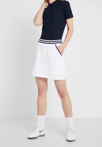Navy polo top with short sleeves and button placket, paired with a white skirt featuring a striped waistband and pockets. White sneakers.