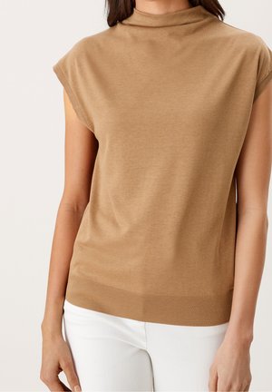 Vrouw die een mouwloze, kamelkleurige gebreide top met een col draagt, gecombineerd met een witte broek, tegen een neutrale achtergrond.