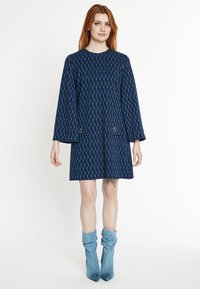 Navyblaues Kleid mit geometrischem Muster, langen Ärmeln und zwei vorderen Taschen. Kombiniert mit hellblauen gefalteten Knöchelstiefeln. Glatte Textur.