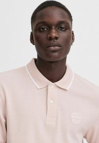 Kortærmet polo shirt i blød pink stof med et tekstureret mønster, hvid krave og et cirkulært logo på brystet.