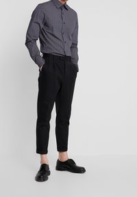 Černá košile s dlouhým rukávem a vzorem, v kombinaci s elegantními černými kalhotami a černými šněrovacími botami. Neutrální pozadí.