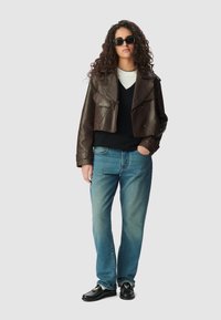 Brązowa skórzana kurtka typu cropped z dwoma przednimi kieszeniami, zestawiona z czarnym swetrem, niebieskimi dżinsami i czarnymi butami loafers z połyskującym wykończeniem.
