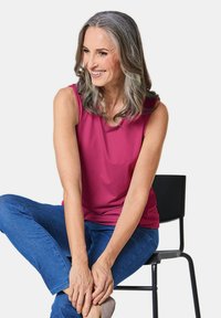 Top senza maniche fucsia con scollatura rotonda, abbinato a jeans blu. Il modello è seduto su una sedia nera, mostrando una postura corporea rilassata.