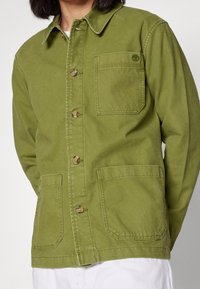 Olivgrön jacka i bomull med krage, framknäppning och två stora framfickor, med en logotypdetalj på bröstet.