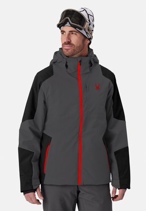 Homme portant une veste de ski grise et noire avec des fermetures éclair rouges, un bonnet blanc à motifs et des lunettes de ski sur le front, regardant sur le côté.