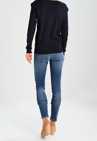 Marineblauer Pullover mit lockerer Passform, kombiniert mit blauen Jeans, die leicht tailliert sind und fransige Säume aufweisen. Neutrale Blockabsatzsandalen runden das Outfit ab.