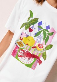 T-shirt blanc présentant un graphique coloré d'une langouste, de citrons, de légumes verts et de vin sur une table à motifs, avec des couleurs vives et des détails.