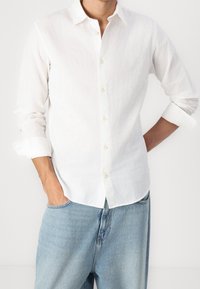 Camicia bianca in lino a maniche lunghe con colletto button-down, caratterizzata da una leggera trama e bottoni beige; abbinata a pantaloni in denim azzurro chiaro.
