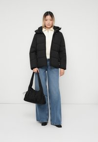 Γυναίκα που φοράει μαύρο μπουφάν, λευκή ζακέτα με φερμουάρ, φαρδιά μπλε τζιν και μαύρες μπότες, κρατώντας μια μαύρη τσάντα ώμου, στέκεται πάνω σε ένα απλό φόντο.