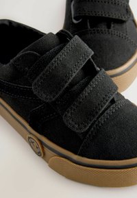 Zwarte canvas schoenen met twee klittenbandbanden, ronde neus en rubberen zool met een gestructureerde gomzool en een logo patch met een vrolijk gezicht.