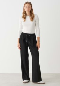 Zwarte, wijde broek met een elastische tailleband en verstelbaar koord, gecombineerd met een langemouwen witte top. Witte sneakers maken de look compleet.
