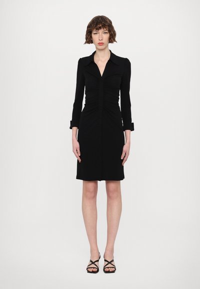 Robe noire avec une silhouette ajustée, des manches longues, un devant à boutons et des détails froncés ; associée à des talons noirs à lanières.