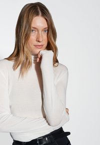 Femme aux cheveux châtains clairs et aux yeux bleus portant un haut à manches longues blanc, reposant son menton sur sa main devant un fond uni.