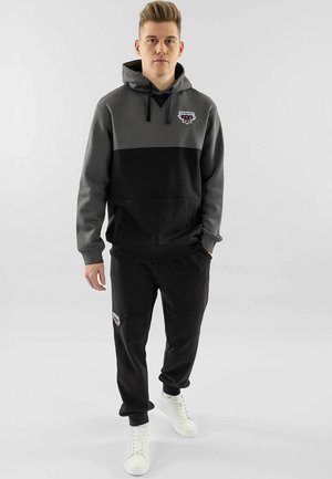 Grauer und schwarzer Kapuzensweatshirt mit Logo auf der Brust. Kombiniert mit schwarzen Jogginghosen und weißen Turnschuhen. Glatte Stofftextur.