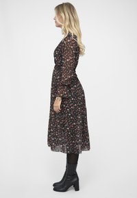 Svart blomstret midi-kjole med gjennomskinnelige lange ermer, prydet med små røde og hvite blomster. Brukt med svarte ankelstøvler. Teksturert stoff.