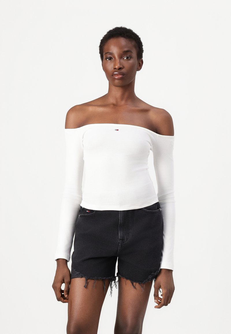 White off-the-shoulder long-sleeve top with a ribbed texture, featuring a small logo detail. Paired with black denim shorts, frayed hem.
