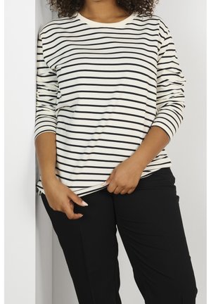 Nainen pukeutuneena pitkähihainen valkoinen paita, jossa mustat vaakarajaukset, ja mustat housut, säätämässä paidan helmaa käsillään.