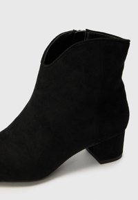 Bottines noires en daim avec une finition texturée, un petit talon carré, et une découpe discrète sur le dessus. Détail zippé pour un enfilage facile.