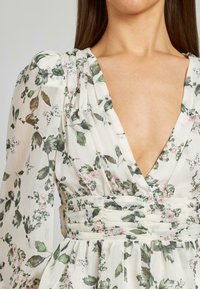 Abito floreale con scollo a V profondo, maniche a sbuffo e vita cinchata. Realizzato in tessuto leggero con uno sfondo crema e motivi verdi, rosa e viola.