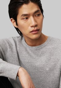 Suéter gris de punto con cuello redondo. Presenta un patrón sutil y un pequeño logo blanco en el pecho. Textura suave, corte holgado.