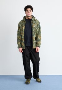 Jachetă din fleece cu model de camuflaj, nuanțe de verde și maro, cu fermoar în față, purtată cu un tricou negru, pantaloni cargo negri și pantofi de drumeție verzi.