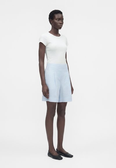 Γυναίκα που στέκεται φορώντας λευκό κοντομάνικο τοπ, ανοιχτό μπλε υφασμάτινη φούστα μέχρι το γόνατο και μαύρα επίπεδα παπούτσια μπροστά σε λευκό απλό φόντο.