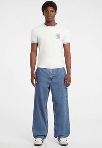 Wit T-shirt met een afbeelding op de borst, gecombineerd met wijde blauwe spijkerbroeken en witte sneakers.